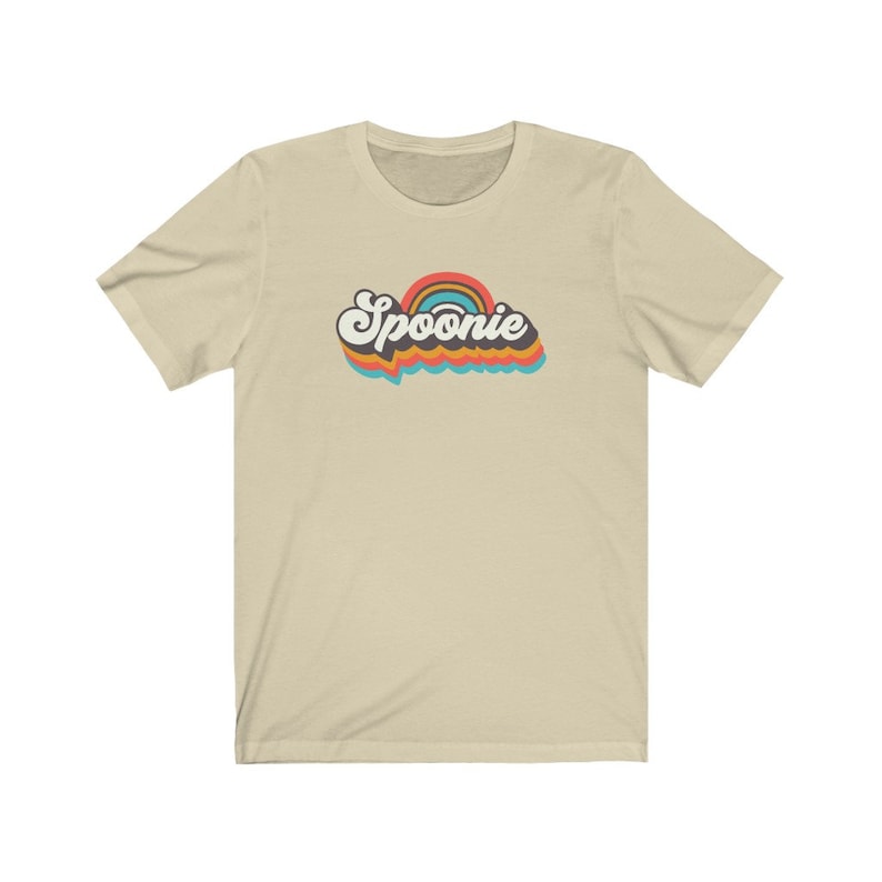 Op de afbeelding: Een beige t-shirt met een retro-regenbooggrafiek en het woord "Spoonie" in zwarte tekst.