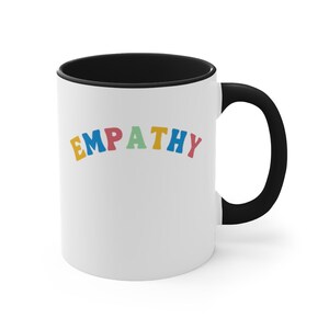 May include: White ceramic coffee mug with a black rim and handle. The mug has the word "EMPATHY" written in rainbow colors.