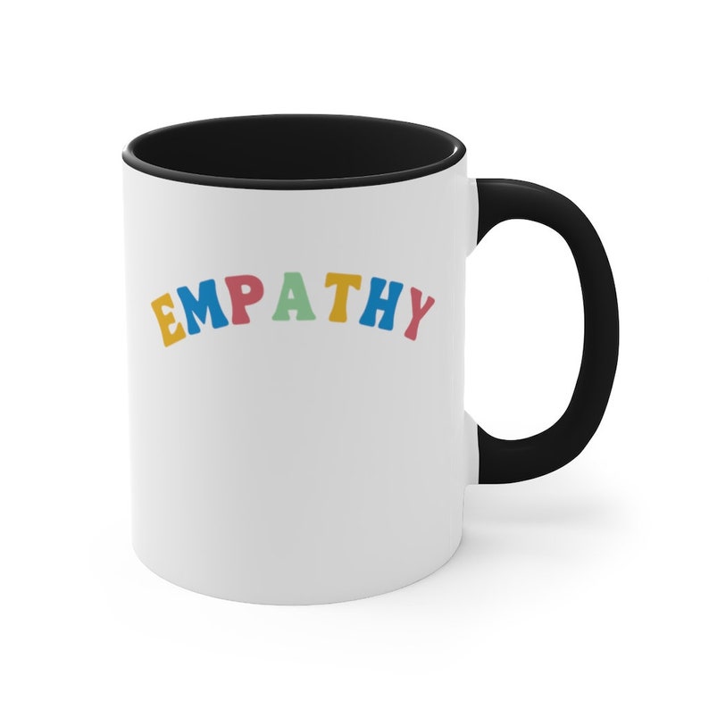 May include: White ceramic coffee mug with a black rim and handle. The mug has the word "EMPATHY" written in rainbow colors.