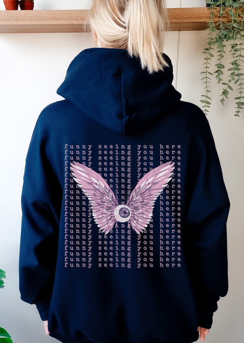 May include: A navy blue hoodie with a pink and white eye with wings on the back. The text "funny seeing you here" is repeated multiple times on the back of the hoodie.