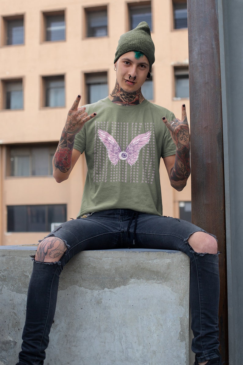 May include: A green t-shirt with a pink eye with wings graphic and the text "Funny seeing you here" repeated multiple times. The person wearing the shirt has green hair and tattoos.