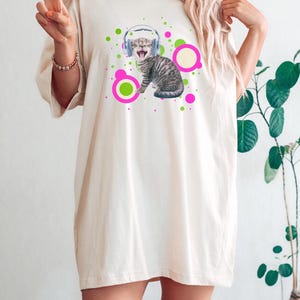 Pu&ograve; includere: T-shirt color crema con grafica di un gatto tigrato grigio con cuffie e circondato da cerchi rosa e verdi. Il gatto ha la bocca aperta. La maglietta ha maniche corte e una vestibilit&agrave; ampia.