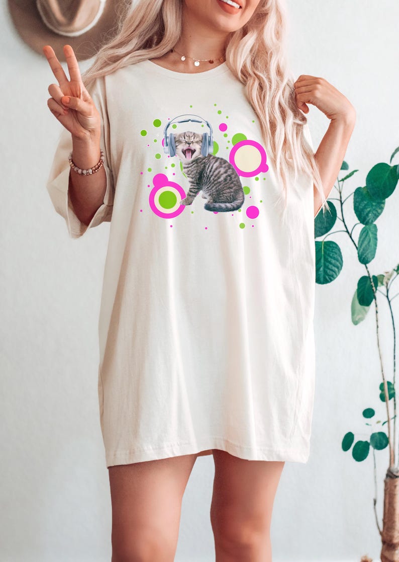 Pu&ograve; includere: T-shirt color crema con grafica di un gatto tigrato grigio con cuffie e circondato da cerchi rosa e verdi. Il gatto ha la bocca aperta. La maglietta ha maniche corte e una vestibilit&agrave; ampia.