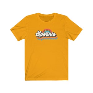 Op de afbeelding: Een geel T-shirt met een retro-stijl grafische afbeelding met de tekst "Spoonie" in een regenboogkleurenschema.
