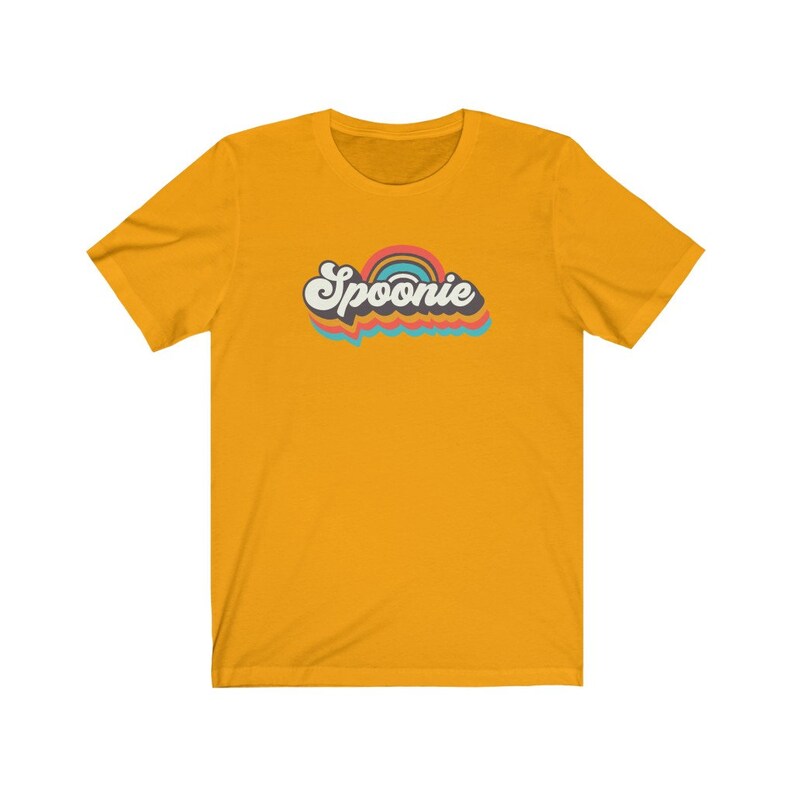 Op de afbeelding: Een geel T-shirt met een retro-stijl grafische afbeelding met de tekst "Spoonie" in een regenboogkleurenschema.