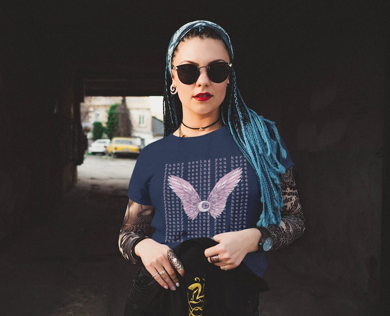 May include: A navy blue t-shirt with a pink and white eye with wings graphic. The text "Funny seeing you here" is repeated in a pattern on the shirt.