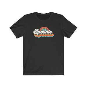 Op de afbeelding: Zwart T-shirt met korte mouwen met een retro regenboogontwerp en het woord "Spoonie" in een kleurrijk, gestileerd lettertype.
