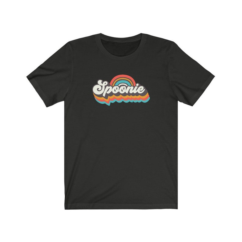 Op de afbeelding: Zwart T-shirt met korte mouwen met een retro regenboogontwerp en het woord "Spoonie" in een kleurrijk, gestileerd lettertype.