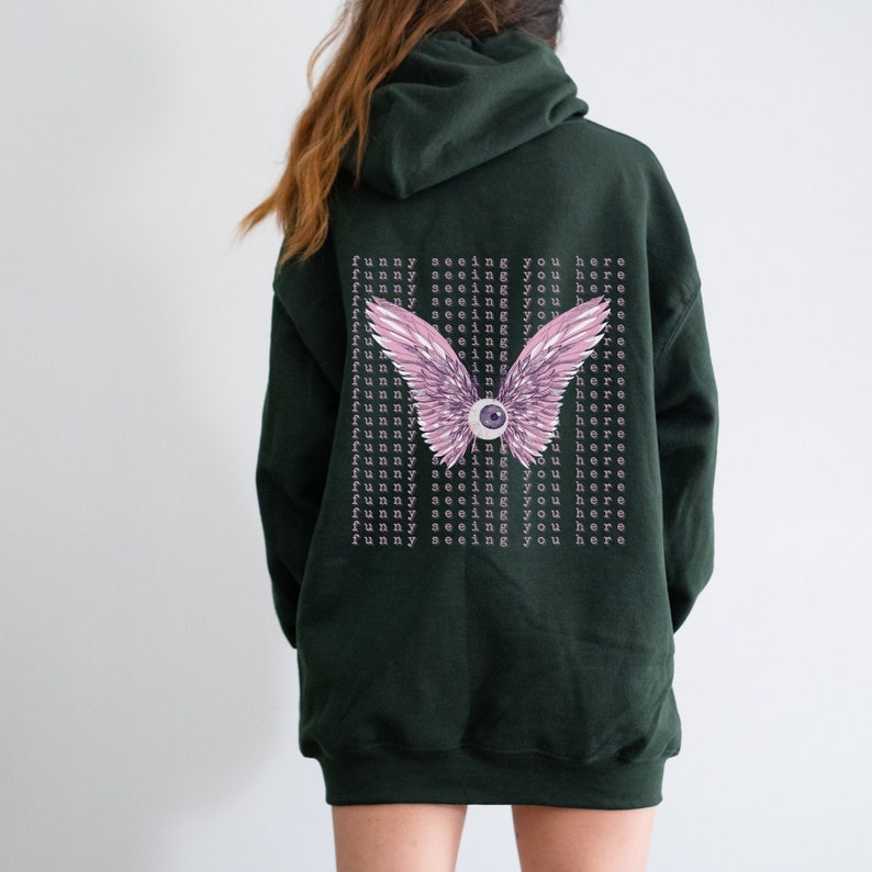 May include: A dark green hooded sweatshirt with a pink and purple eye with wings on the back. The text "funny seeing you here" is repeated multiple times on the back of the sweatshirt.