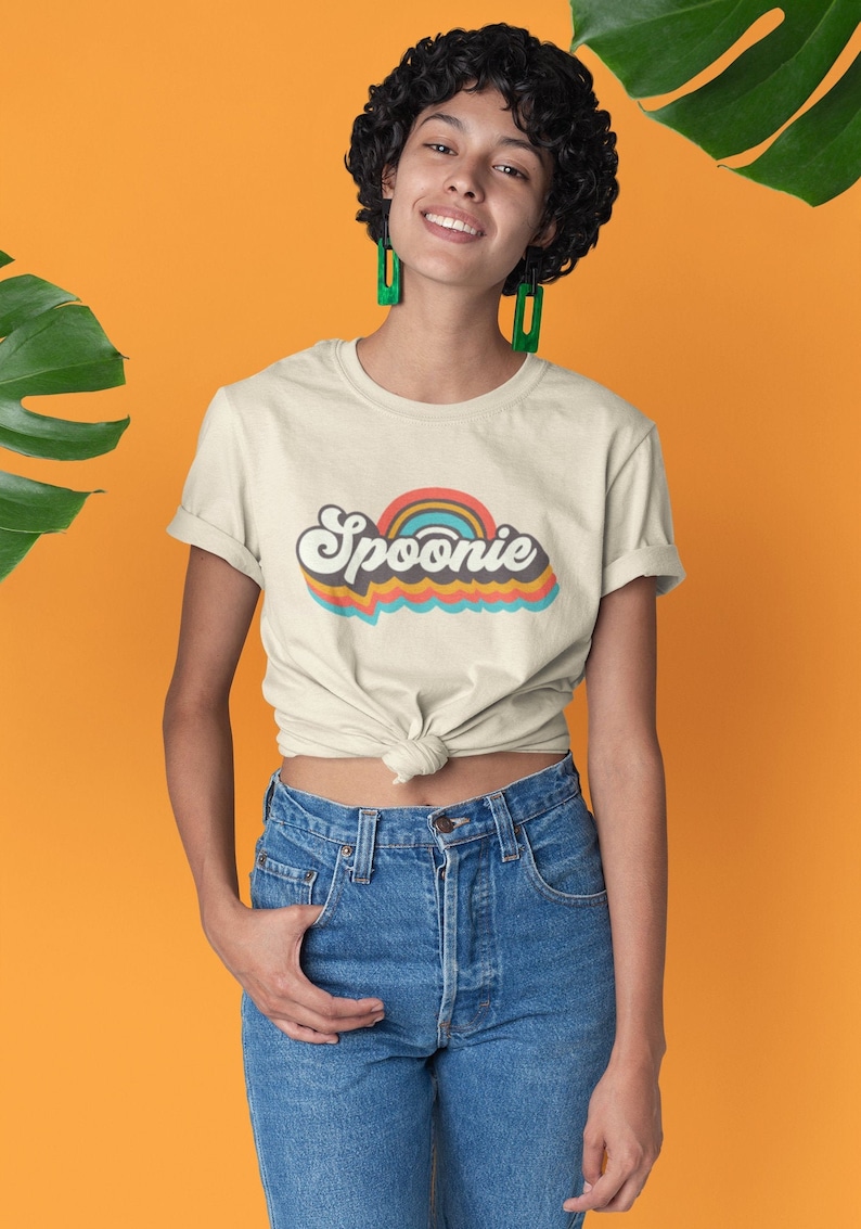 Op de afbeelding: Een persoon draagt een lichtbeige t-shirt met een retro regenbooggrafiek en de tekst "Spoonie" in een kleurrijk lettertype. De persoon draagt een blauwe jeans en heeft kort krullend haar.
