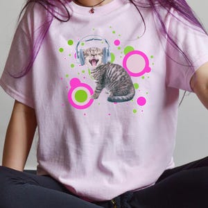 Pu&ograve; includere: T-shirt rosa chiaro con una grafica di un gatto tigrato con le cuffie e la bocca aperta. Il design include cerchi verdi e rosa. La maglietta &egrave; a maniche corte.