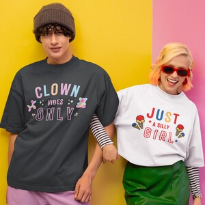 May include: Two people wearing t-shirts with colorful graphic designs. The person on the left is wearing a black t-shirt with the text "Clown Vibes Only" in rainbow colors. The person on the right is wearing a white t-shirt with the text "Just a Silly Girl" in red, blue, and yellow colors.