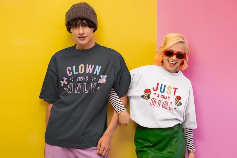 May include: Two people wearing t-shirts with colorful graphic designs. The person on the left is wearing a black t-shirt with the text "Clown Vibes Only" in rainbow colors. The person on the right is wearing a white t-shirt with the text "Just a Silly Girl" in red, blue, and yellow colors.
