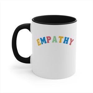 May include: White ceramic coffee mug with a black rim and handle. The mug has the word "EMPATHY" written in rainbow colors.