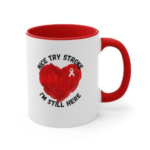 May include: White ceramic coffee mug with a red rim and handle. The mug has a red heart with a white ribbon on it and the text "Nice try stroke I'm still here" around it.