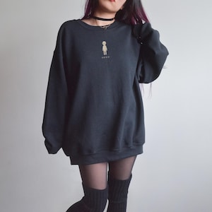 May include: A black crewneck sweatshirt featuring a small, centered beige graphic of a stylized alien figure above the word "SOON" in a simple, sans-serif font. The garment has long sleeves, a ribbed neckline, and a relaxed, oversized fit.