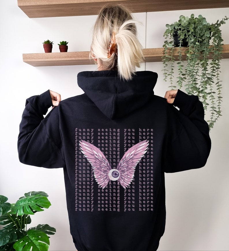 May include: Black hooded sweatshirt with a pink and purple eye with wings graphic on the back. The text "Funny seeing you here" is repeated multiple times on the back of the sweatshirt.