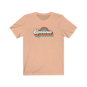Op de afbeelding: Een perzikkleurig T-shirt met een retro regenboogontwerp en het woord "Spoonie" in zwarte tekst.