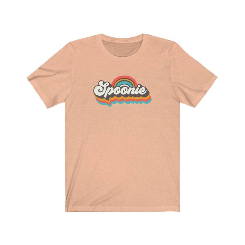 Op de afbeelding: Een perzikkleurig T-shirt met een retro regenboogontwerp en het woord "Spoonie" in zwarte tekst.
