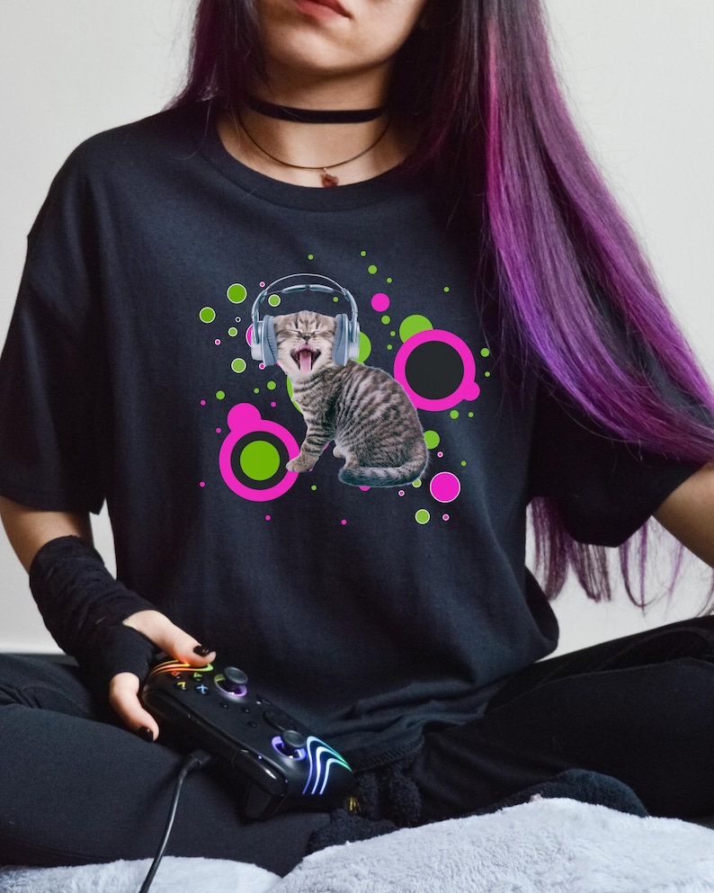 Pu&ograve; includere: T-shirt nero con un gatto tigrato con le cuffie e la bocca spalancata, circondato da cerchi rosa e verdi. La persona tiene in mano un controller di gioco nero. La maglietta &egrave; in stile casual.