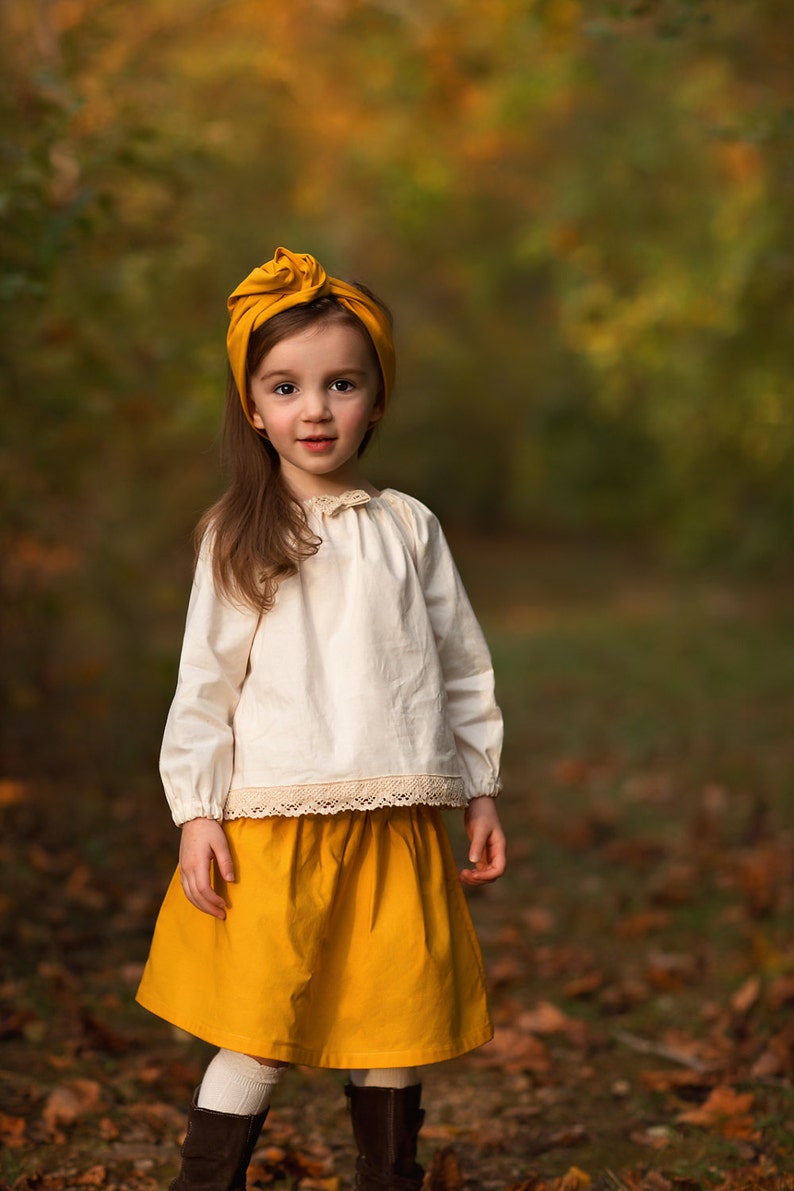 May include: A young child wearing a mustard yellow headband, a cream-colored long-sleeved blouse with lace trim, and a matching mustard yellow skirt. The child is also wearing white socks and brown boots. The background is a blurred autumn scene.
