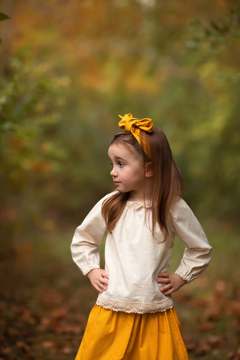 May include: A young person wearing a mustard yellow headband and a matching skirt. The outfit includes a cream-colored long-sleeved blouse with lace trim. The background is a blurred view of a forest with autumn colors.