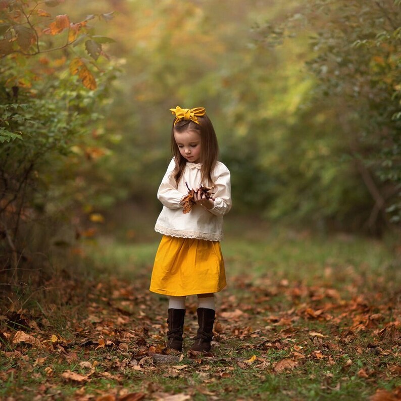 May include: A young person in a white blouse with lace trim and a yellow skirt stands in a forest path. They wear brown boots and a yellow bow in their hair, holding fallen leaves. The scene is surrounded by autumn foliage.