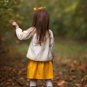 May include: A child in a cream-colored blouse with lace trim and a mustard yellow skirt points towards the right. The child has long brown hair, a yellow bow, white socks, and brown boots. The background is a blurred forest scene with fallen leaves.