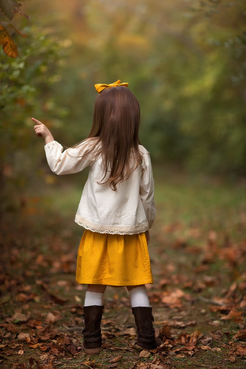 May include: A child in a cream-colored blouse with lace trim and a mustard yellow skirt points towards the right. The child has long brown hair, a yellow bow, white socks, and brown boots. The background is a blurred forest scene with fallen leaves.