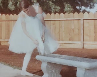 May include: A person wearing a white ballet tutu and white tights stands on a concrete path next to a light gray stone bench. The person is leaning forward while adjusting a white ballet slipper on their foot in an outdoor setting.