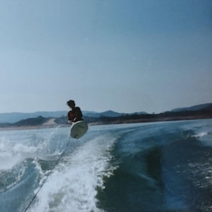 May include: A person wakeboarding on a large body of water, riding a white board while being pulled by a tow rope. The scene features a prominent white wake trail behind a boat, with distant hills visible under a clear blue sky during the day.