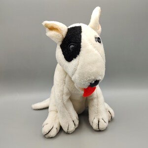 May include: A plush white Bull Terrier dog with a black patch over one eye, black nose, and a red tongue. The stuffed animal is sitting upright with its paws forward, and its ears are perked up.