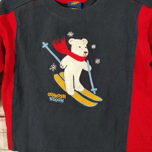 May include: A navy blue long-sleeved shirt with red side panels. The shirt features an embroidered image of a white polar bear wearing a red scarf and skiing on yellow skis. The text "OSHKOSH B'GOSH" is printed on the shirt.