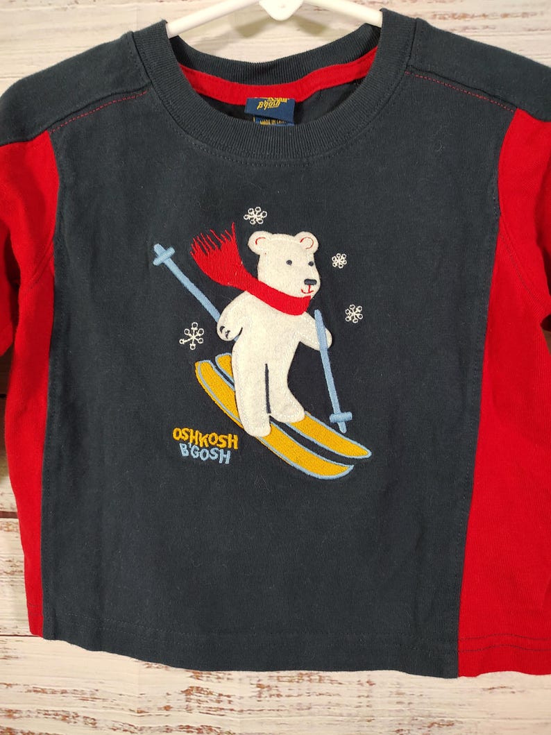 May include: A navy blue long-sleeved shirt with red side panels. The shirt features an embroidered image of a white polar bear wearing a red scarf and skiing on yellow skis. The text "OSHKOSH B'GOSH" is printed on the shirt.