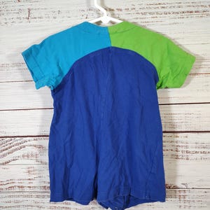 May include: A colorful romper in shades of blue and green, hanging on a white hanger. The top is divided into sections of turquoise and lime green, with a deep blue lower section. The garment has short sleeves and a round neckline.