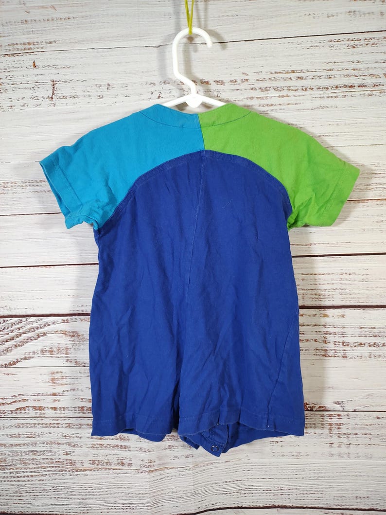 May include: A colorful romper in shades of blue and green, hanging on a white hanger. The top is divided into sections of turquoise and lime green, with a deep blue lower section. The garment has short sleeves and a round neckline.