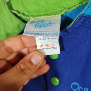 May include: Close-up of a blue and green baby's shirt with a tag that reads "Baby B'gosh". The shirt has green buttons and the text "Ocean seahorse and" is visible. The tag indicates the garment is for 18 months.