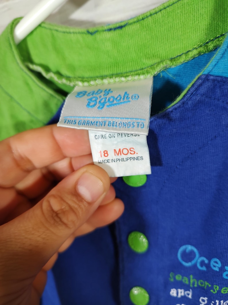 May include: Close-up of a blue and green baby's shirt with a tag that reads "Baby B'gosh". The shirt has green buttons and the text "Ocean seahorse and" is visible. The tag indicates the garment is for 18 months.