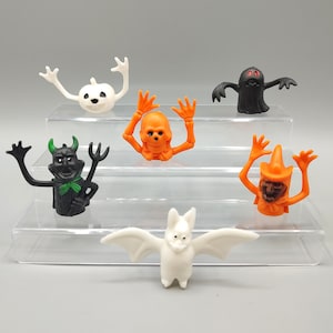 May include: A collection of Halloween-themed figurines, including a white pumpkin, a black ghost, an orange skull, a black devil, an orange witch, and a white bat. The figurines are arranged on clear, tiered display shelves.