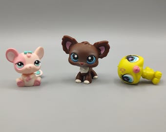 Puede incluir: Tres pequeñas figuras de animales de plástico de colores. Una figura rosa y blanca con forma de ratón con un lazo azul, una figura marrón con forma de perro con ojos azules y una figura amarilla con forma de pollito con mejillas rosas.