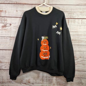 May include: Black sweatshirt with a cream-colored collar and cuffs. The front features an applique of three stacked orange jack-o'-lanterns with smiling faces. Yellow stars and bats are also appliqued on the front, creating a Halloween theme.