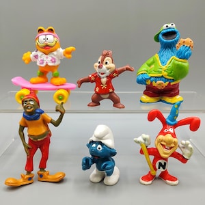 May include: A collection of colorful cartoon character figurines, including Garfield on a skateboard, Chip, Cookie Monster, a Smurf, and a red and yellow jester-like figure. The figures are made of plastic and are posed in various stances.
