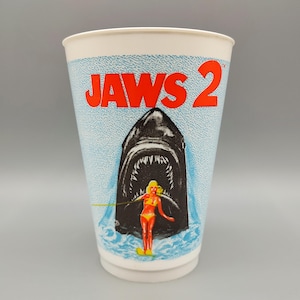 May include: A white paper cup featuring the movie title "JAWS 2" in red letters. The cup has a blue water pattern and a graphic of a shark with an open mouth and a woman in a yellow bikini. The cup is a vintage collectible.