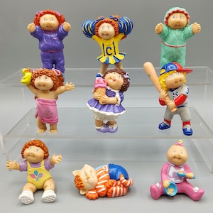 May include: A collection of vintage Cabbage Patch Kids figurines. The dolls are in various outfits, including a cheerleader, a baseball player, and a baby in a pink outfit. The figurines are made of plastic and are brightly colored.