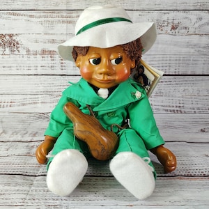 May include: A green and white doll with a white hat and a wooden horn. The doll is sitting on a white wooden surface.
