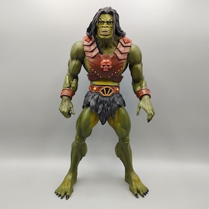 May include: A green action figure of a muscular humanoid with long black hair. The figure wears a brown leather chest piece with a skull emblem, a fur loincloth, and brown wristbands. The figure has a menacing expression.