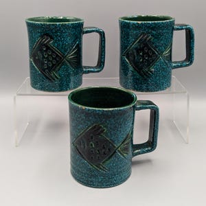 May include: Three teal blue ceramic mugs with black fish designs. The mugs have a speckled glaze and a rounded handle.