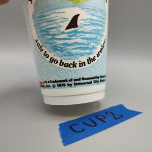 May include: A white paper cup with a blue and white ocean design, featuring a shark fin. Text on the cup reads "safe to go back in the water." A blue tape with "CUP2" written on it is in the foreground.