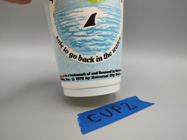 May include: A white paper cup with a blue and white ocean design, featuring a shark fin. Text on the cup reads "safe to go back in the water." A blue tape with "CUP2" written on it is in the foreground.