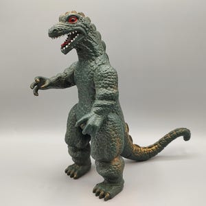 May include: A green and gold Godzilla-like toy figure with red eyes and an open mouth. The figure has a textured, scaly appearance and is posed with one arm extended. The tail curves to the right. The toy is made of plastic.
